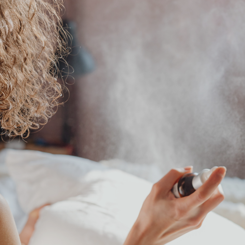 A close-up view of a person with curly hair spraying a mist from a bottle onto a white pillow on a bed. Soft background with a hint of a lamp.