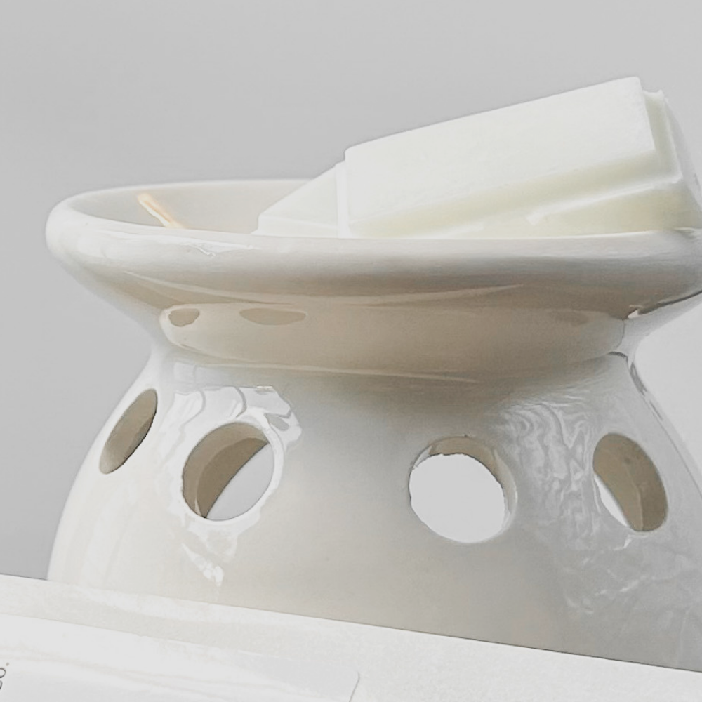Close-up of a white ceramic wax warmer with an unlit candle in the bowl. The warmer features holes for ventilation and is set against a neutral background.