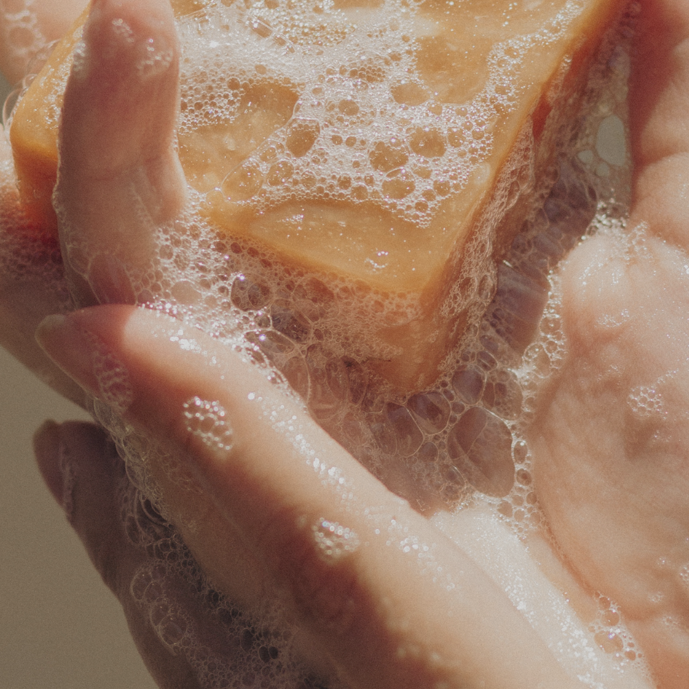 Close-up of a hand holding a bar of soap with bubbles forming around it, indicating washing action. The soap is light brown, surrounded by foamy suds.