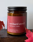 An amber glass jar candle with a black lid reading 'orchard stroll apple butter + maple' by Cedar Moon Candle Co., styled on a dark wooden surface with red autumn leaves against a light gray background.