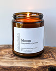 A lit amber jar candle labeled 'bloom honeysuckle jasmine' sits on a wooden surface. The candle is made from a cocowax blend and was hand poured in Oregon. The flame gently flickers.