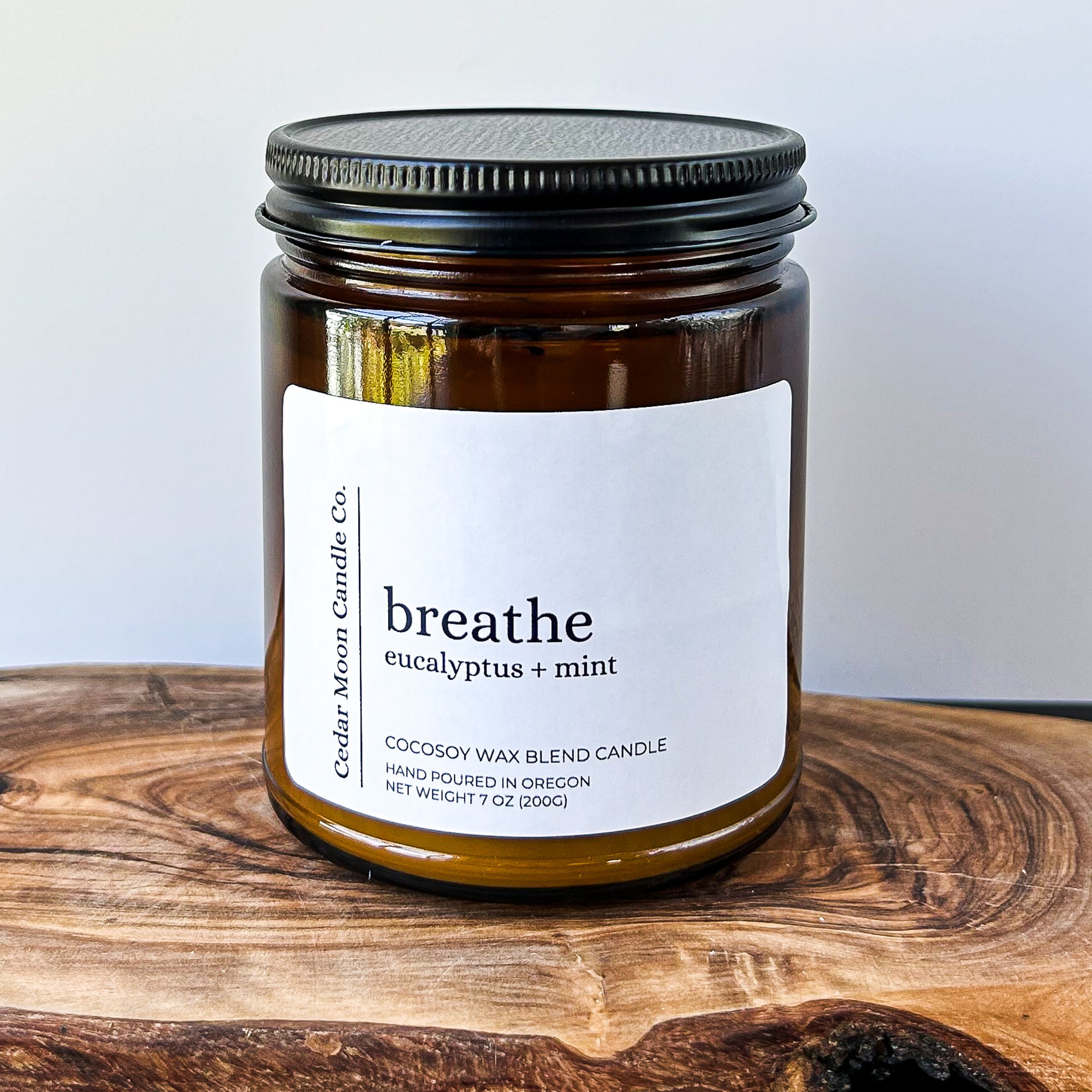 A close-up of a brown glass candle jar labeled 'breathe' with scents of eucalyptus and mint, placed on a wooden surface. The candle is a 7 oz cocowax blend, hand poured in Oregon.