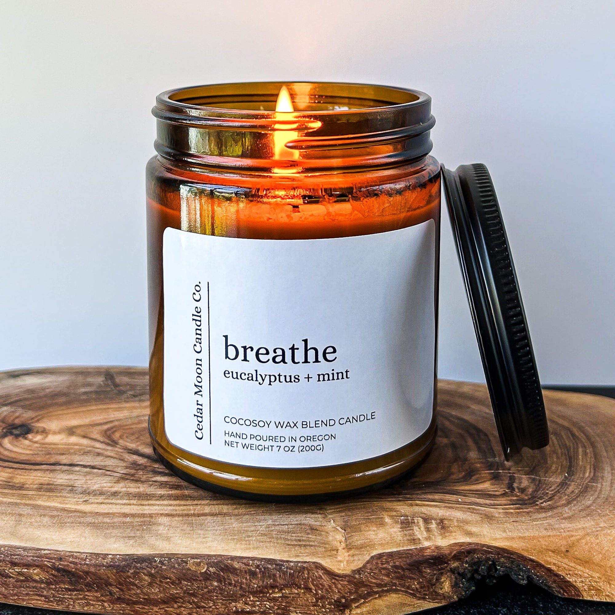 Close-up of a burning candle in a glass jar labeled 'breathe' with eucalyptus and mint scent, sitting on a wooden surface with a black lid beside it.
