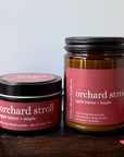 Two amber glass and metal candles from Cedar Moon Candle Co. labeled 'orchard stroll apple butter + maple,' displayed on a dark wooden surface with red autumn leaves against a light gray background.