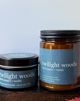 Two Twilight Woods scented candles, one in a round tin and one in a glass jar, displayed on a dark wooden surface with autumn leaves.