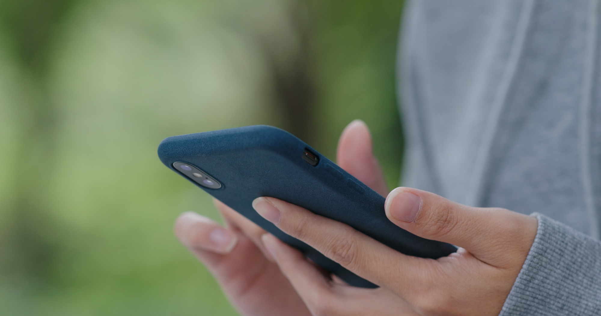 Close-up of a person holding a smartphone in a blue case, outdoors with a blurred green background. The person's fingers are gently gripping the phone.