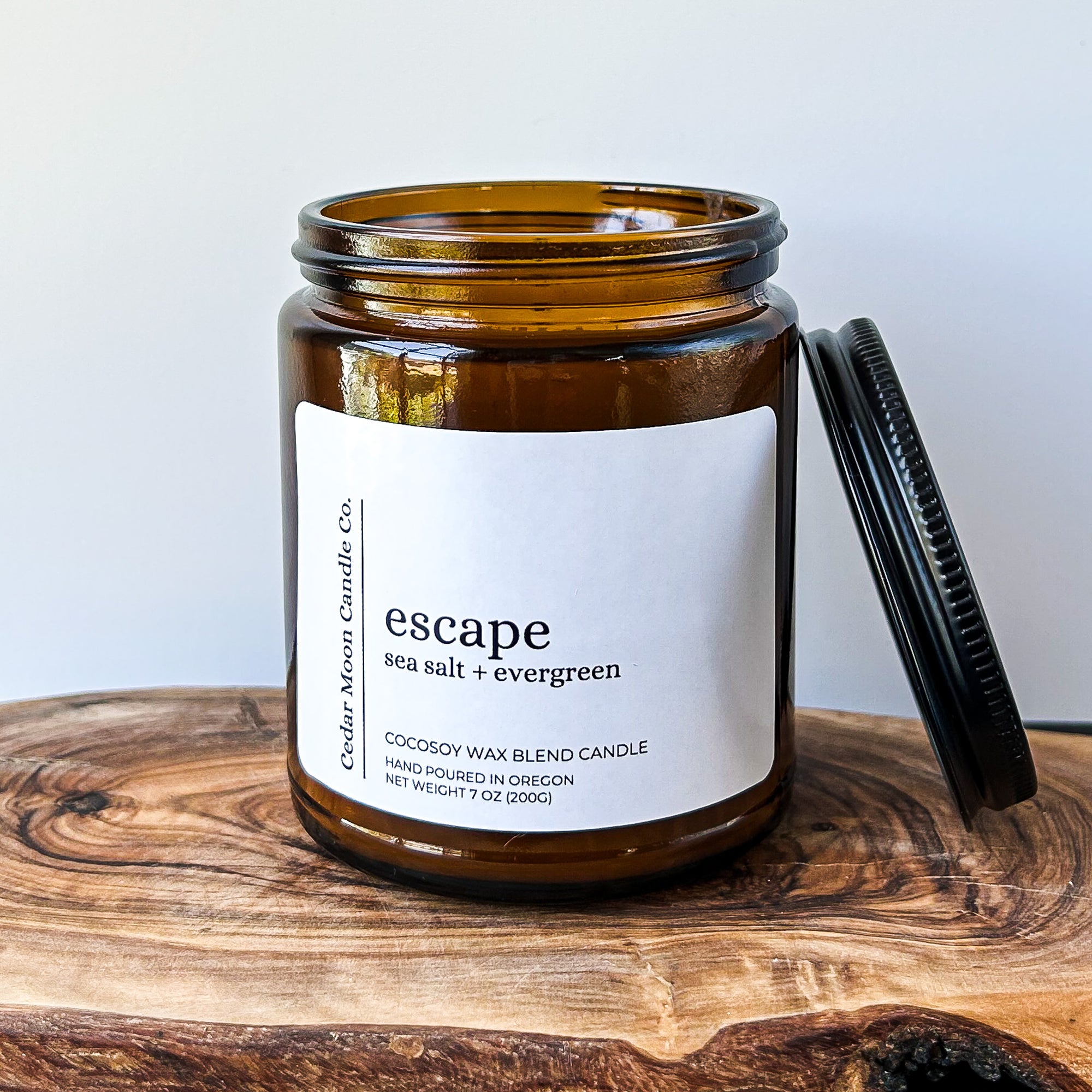 A brown glass candle jar labeled 'escape', featuring a sea salt and evergreen scent. The jar is sitting on a wooden surface with a black lid positioned next to it.