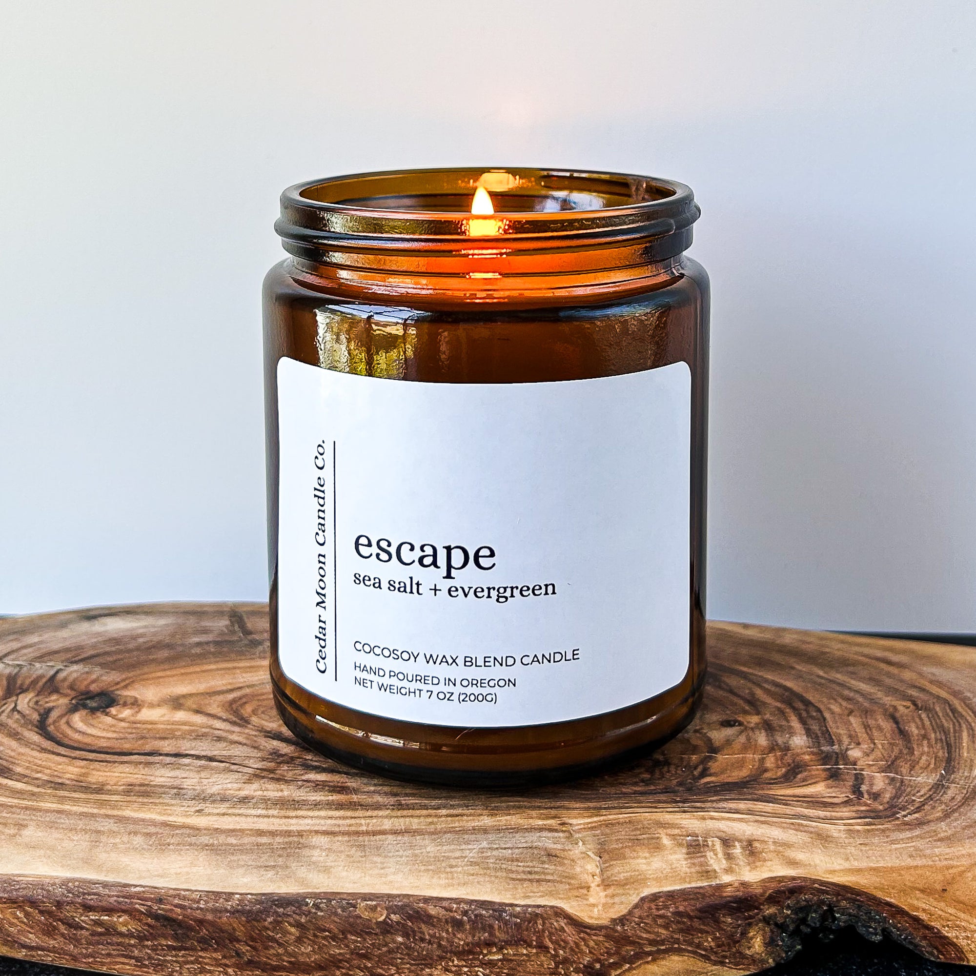 A lit amber glass candle labeled 'escape sea salt evergreen' on a wooden surface. The candle is made of a cocosoy wax blend and is hand poured in Oregon.