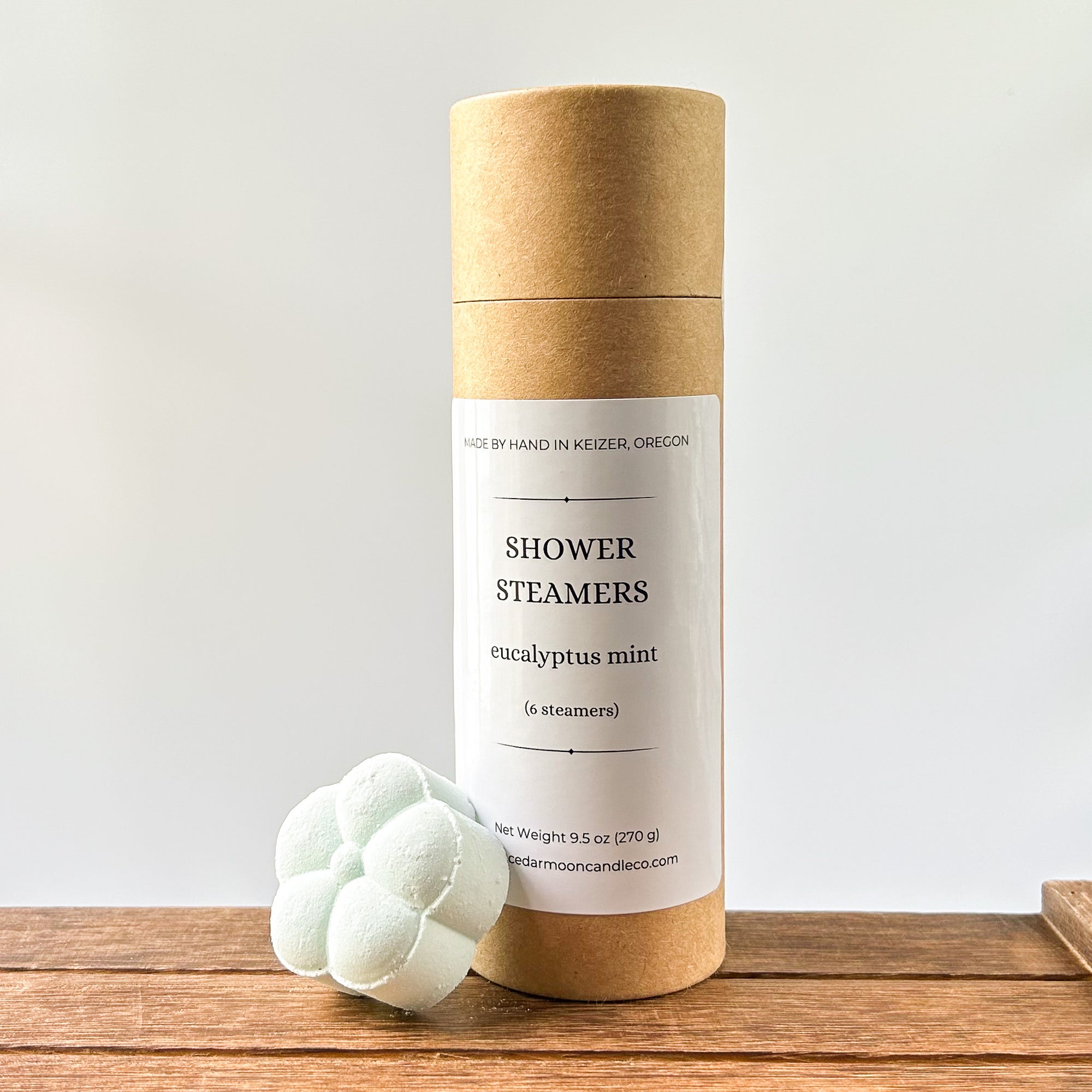 A cylindrical cardboard package labeled 'Shower Steamers' with a eucalyptus mint scent, featuring a white label. One light mint colored shower steamer is placed beside the package on a wooden surface.