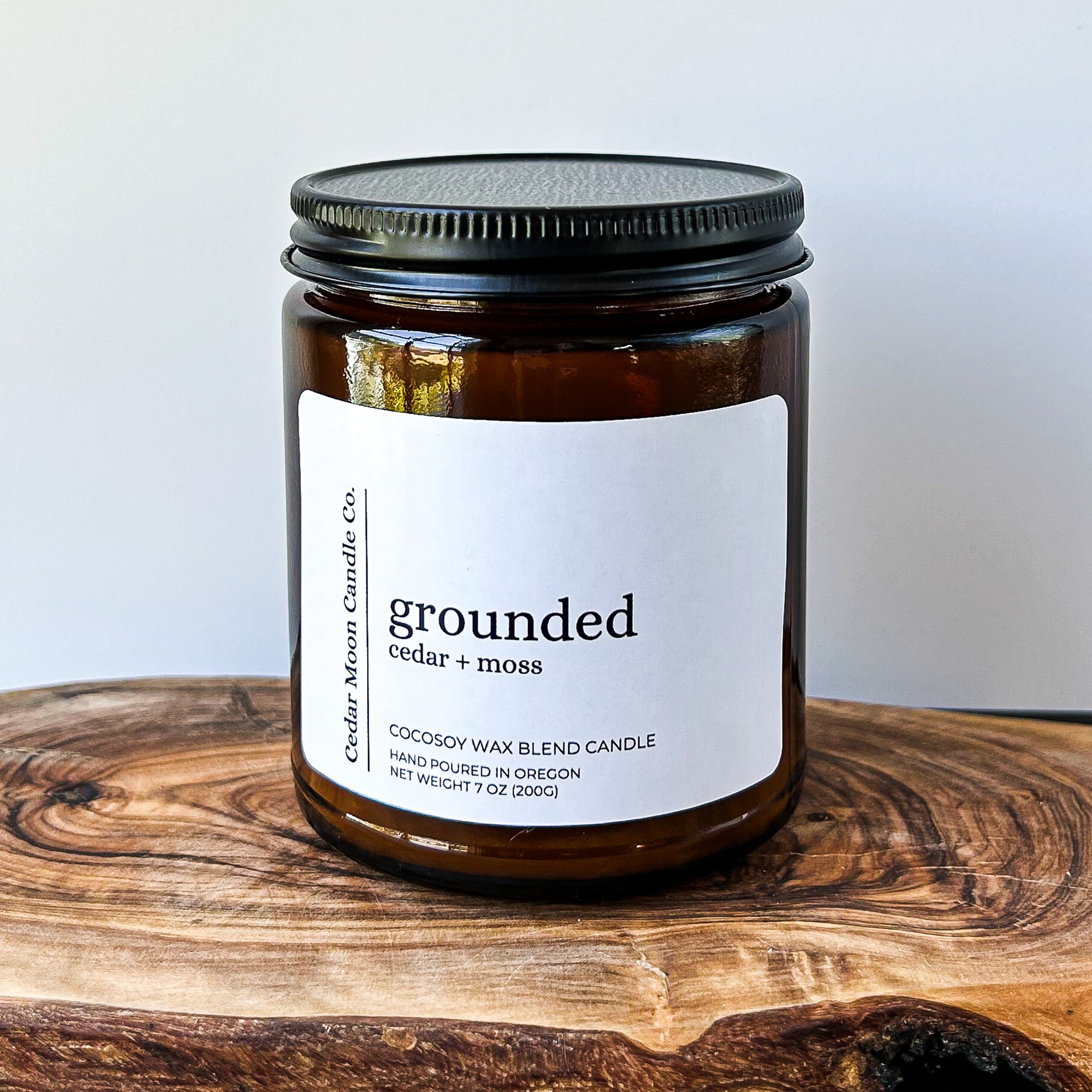 A close-up of a brown glass candle jar labeled 'grounded' with scents of cedar and moss, placed on a wooden surface. The candle is a 7 oz cocowax blend, hand poured in Oregon.