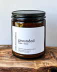 A close-up of a brown glass candle jar labeled 'grounded' with scents of cedar and moss, placed on a wooden surface. The candle is a 7 oz cocowax blend, hand poured in Oregon.