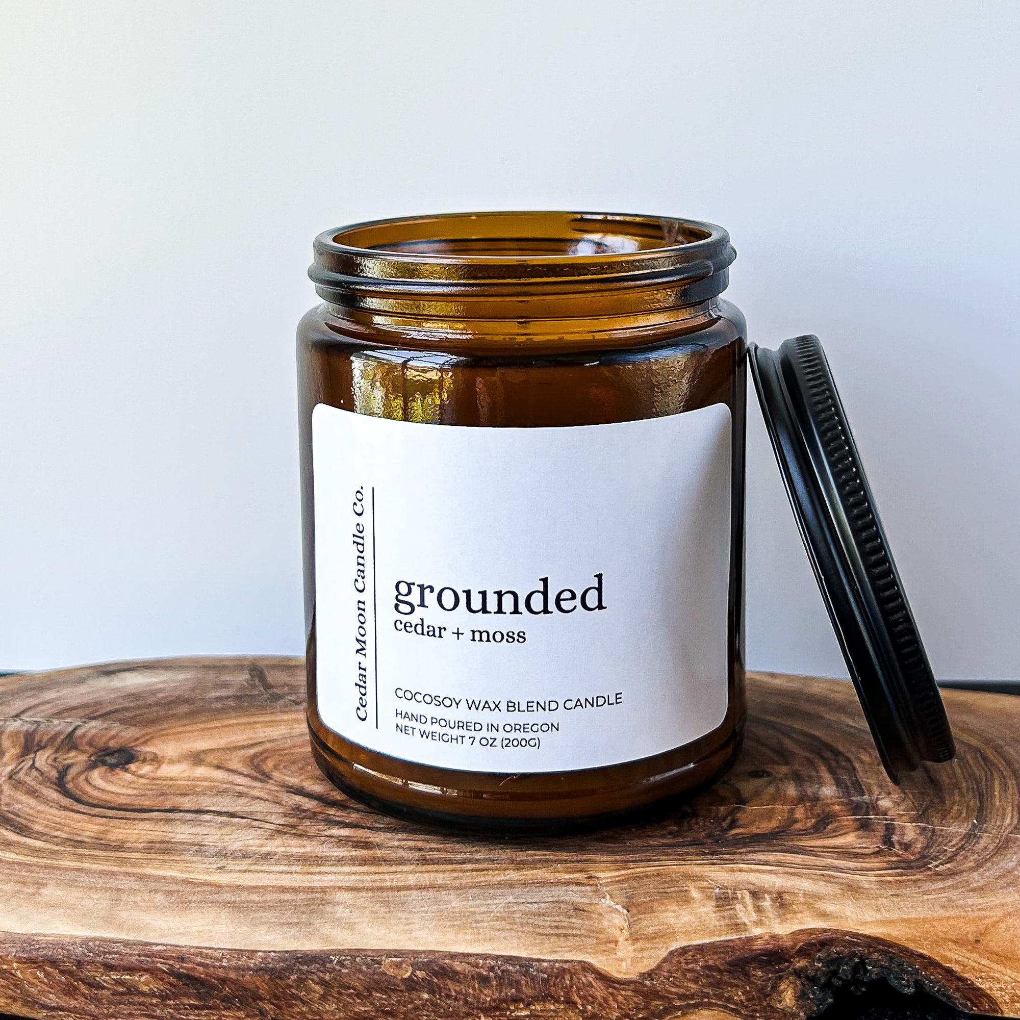A brown glass candle jar labeled 'grounded', featuring a cedar and moss scent. The jar is sitting on a wooden surface with a black lid positioned next to it.
