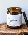 A brown glass candle jar labeled 'grounded', featuring a cedar and moss scent. The jar is sitting on a wooden surface with a black lid positioned next to it.