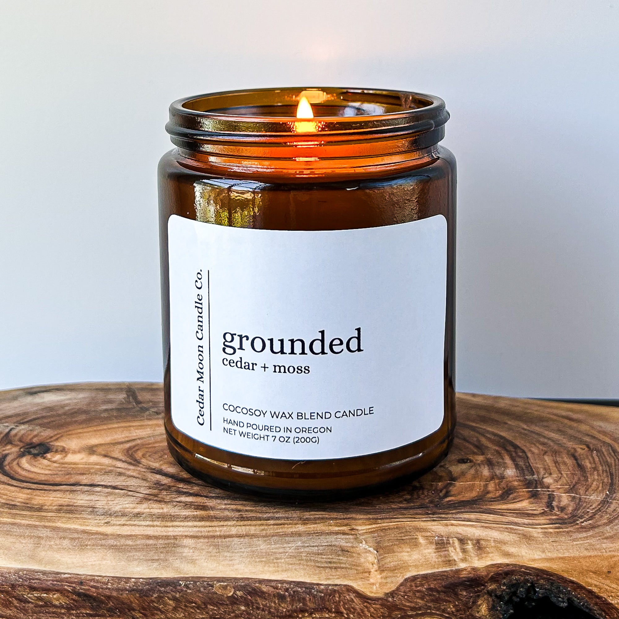 A lit amber glass candle labeled 'grounded cedar and moss' on a wooden surface. The candle is made of a cocosoy wax blend and is hand poured in Oregon.