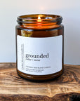 A lit amber glass candle labeled 'grounded cedar and moss' on a wooden surface. The candle is made of a cocosoy wax blend and is hand poured in Oregon.