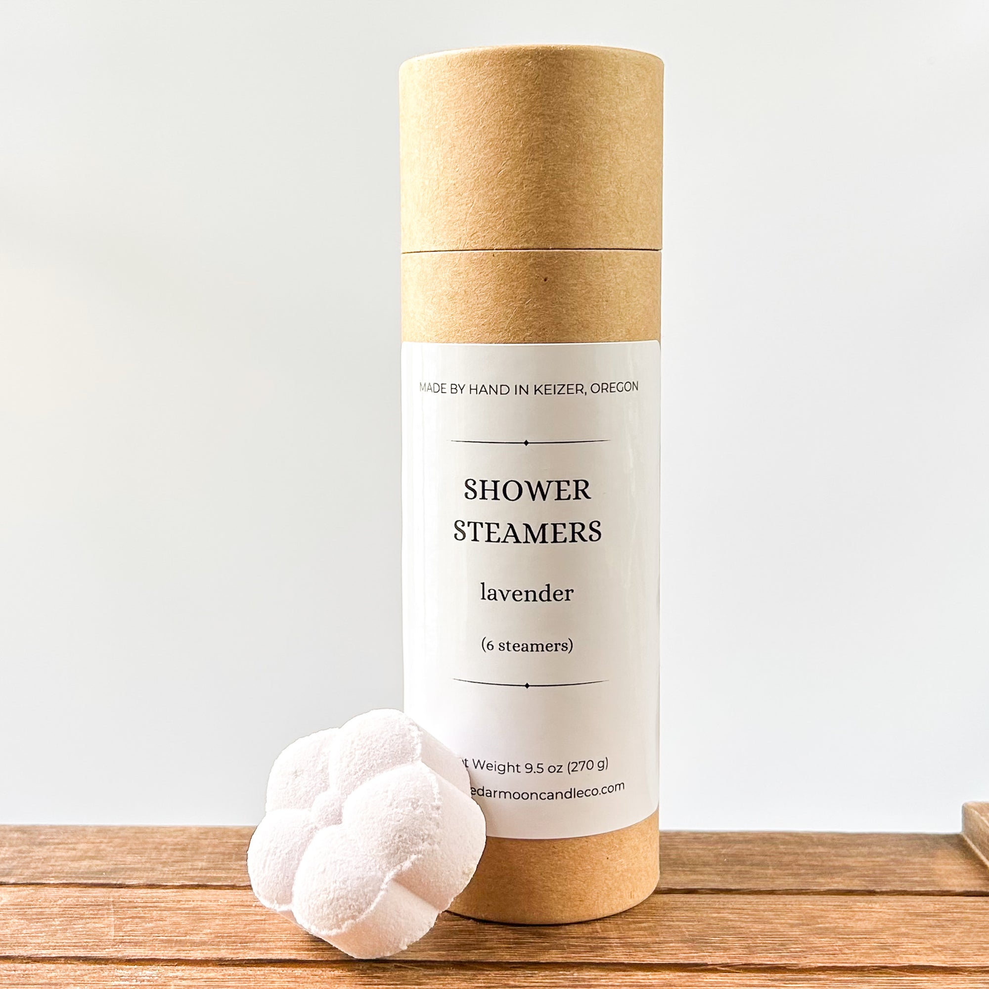 A cylindrical cardboard package labeled 'Shower Steamers' with a lavender scent, featuring a white label. One light lavender colored shower steamer is placed beside the package on a wooden surface.