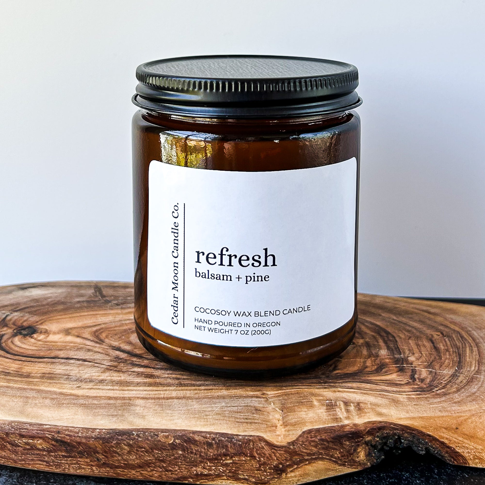 A close-up of a brown glass candle jar labeled 'refresh' with scents of balsam and pine, placed on a wooden surface. The candle is a 7 oz cocowax blend, hand poured in Oregon.