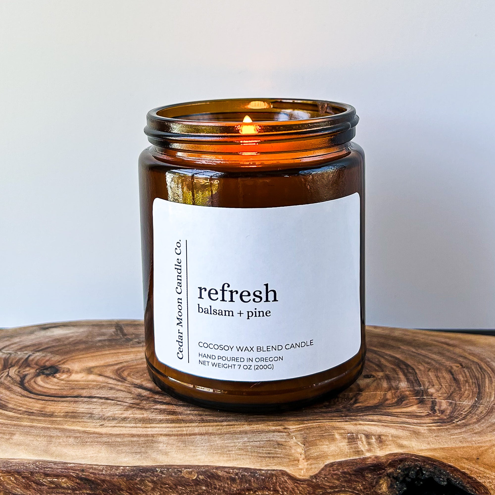 A lit amber glass candle labeled 'refresh balsam and pine' on a wooden surface. The candle is made of a cocosoy wax blend and is hand poured in Oregon.