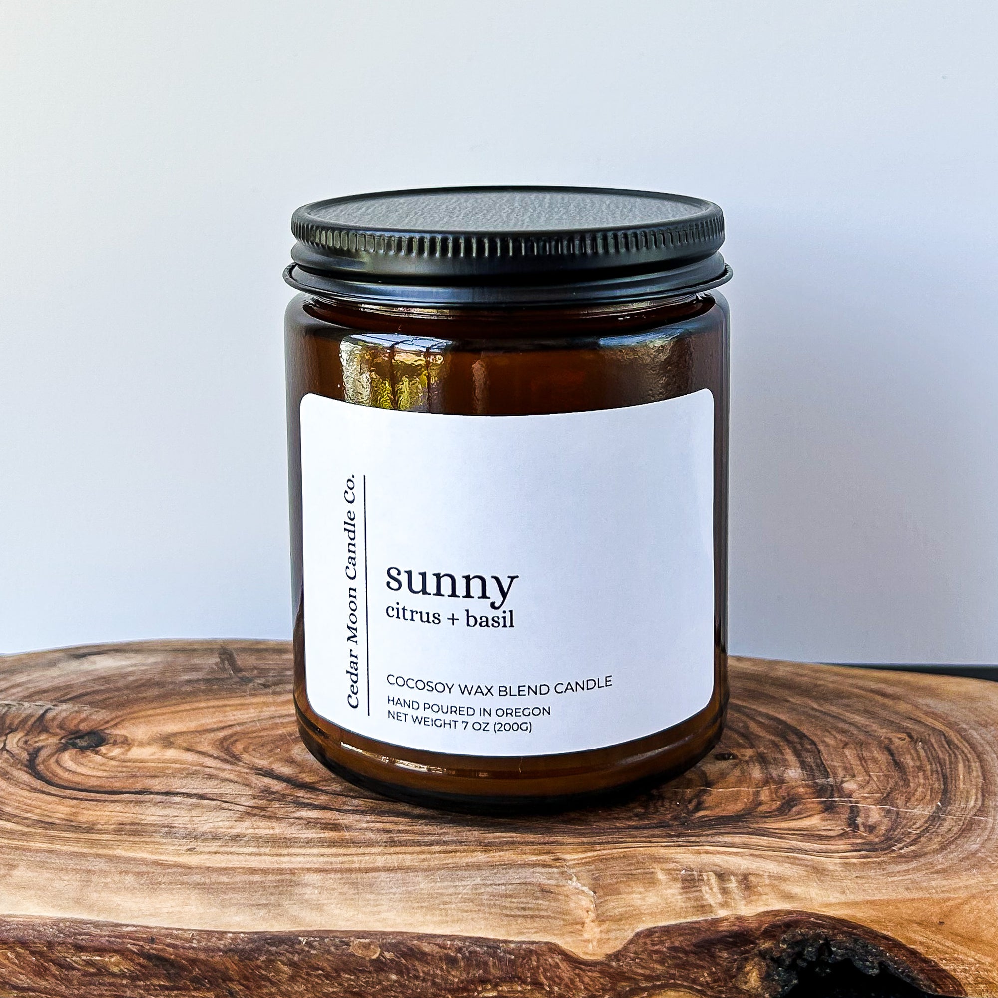 A close-up of a brown glass candle jar labeled 'sunny' with scents of citrus and basil, placed on a wooden surface. The candle is a 7 oz cocowax blend, hand poured in Oregon.