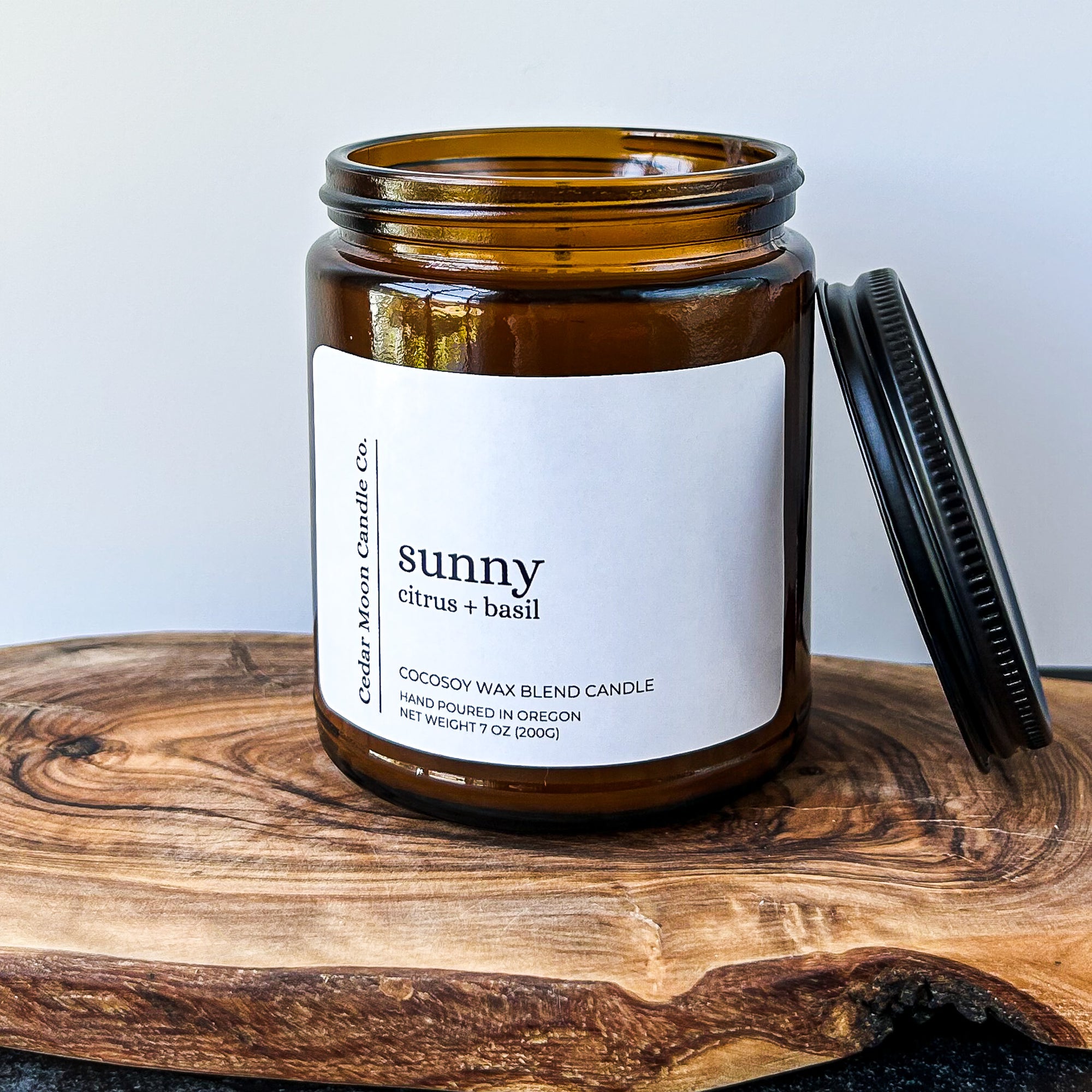 A brown glass candle jar labeled 'sunny', featuring a citrus and basil scent. The jar is sitting on a wooden surface with a black lid positioned next to it.