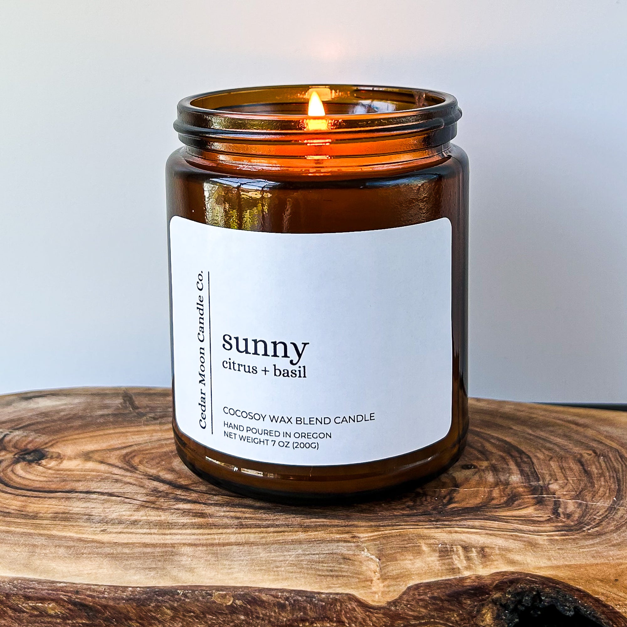 A lit amber glass candle labeled 'sunny citrus and basil' on a wooden surface. The candle is made of a cocosoy wax blend and is hand poured in Oregon.