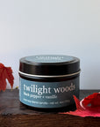 A black candle tin labeled 'twilight woods black pepper + vanille' sits on a dark wooden surface with red autumn leaves around it against a plain light background.