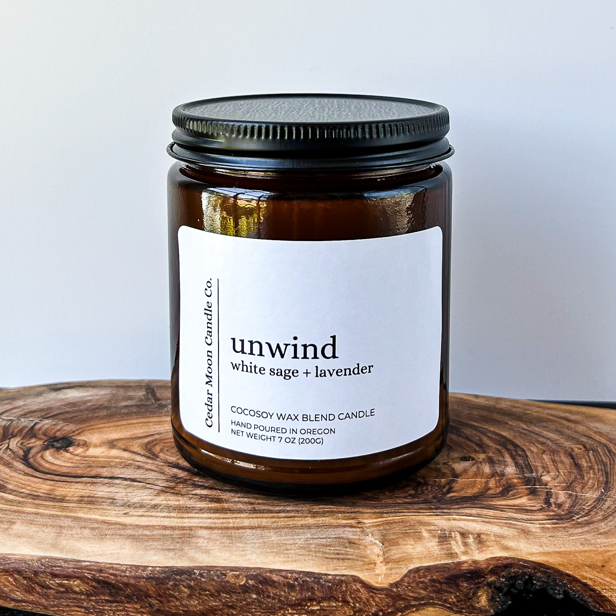 A close-up of a brown glass candle jar labeled 'sunny' with scents of citrus and basil, placed on a wooden surface. The candle is a 7 oz cocosoywax blend, hand poured in Oregon.