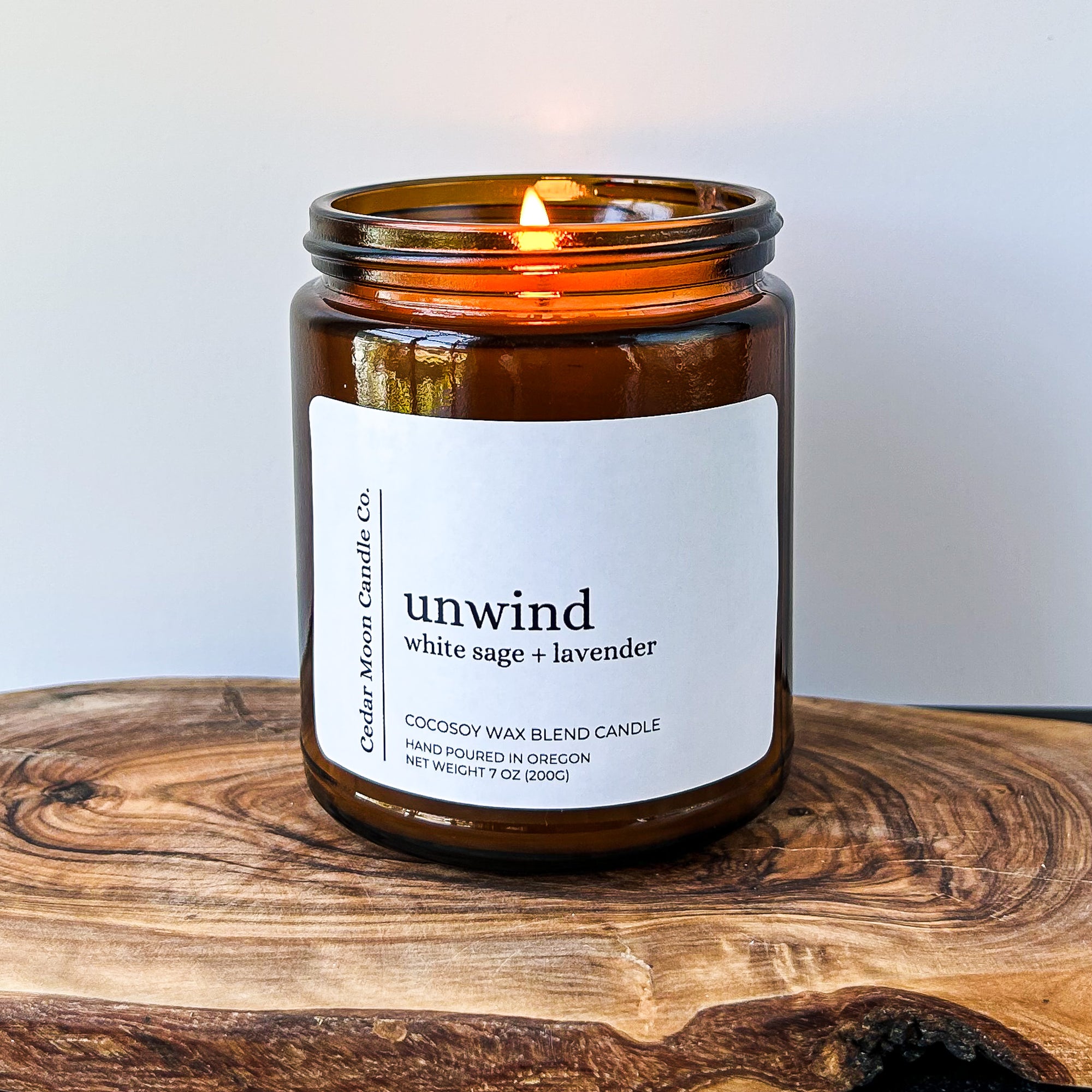 A lit amber glass candle labeled 'unwind white sage and lavender' on a wooden surface. The candle is made of a cocosoy wax blend and is hand poured in Oregon.