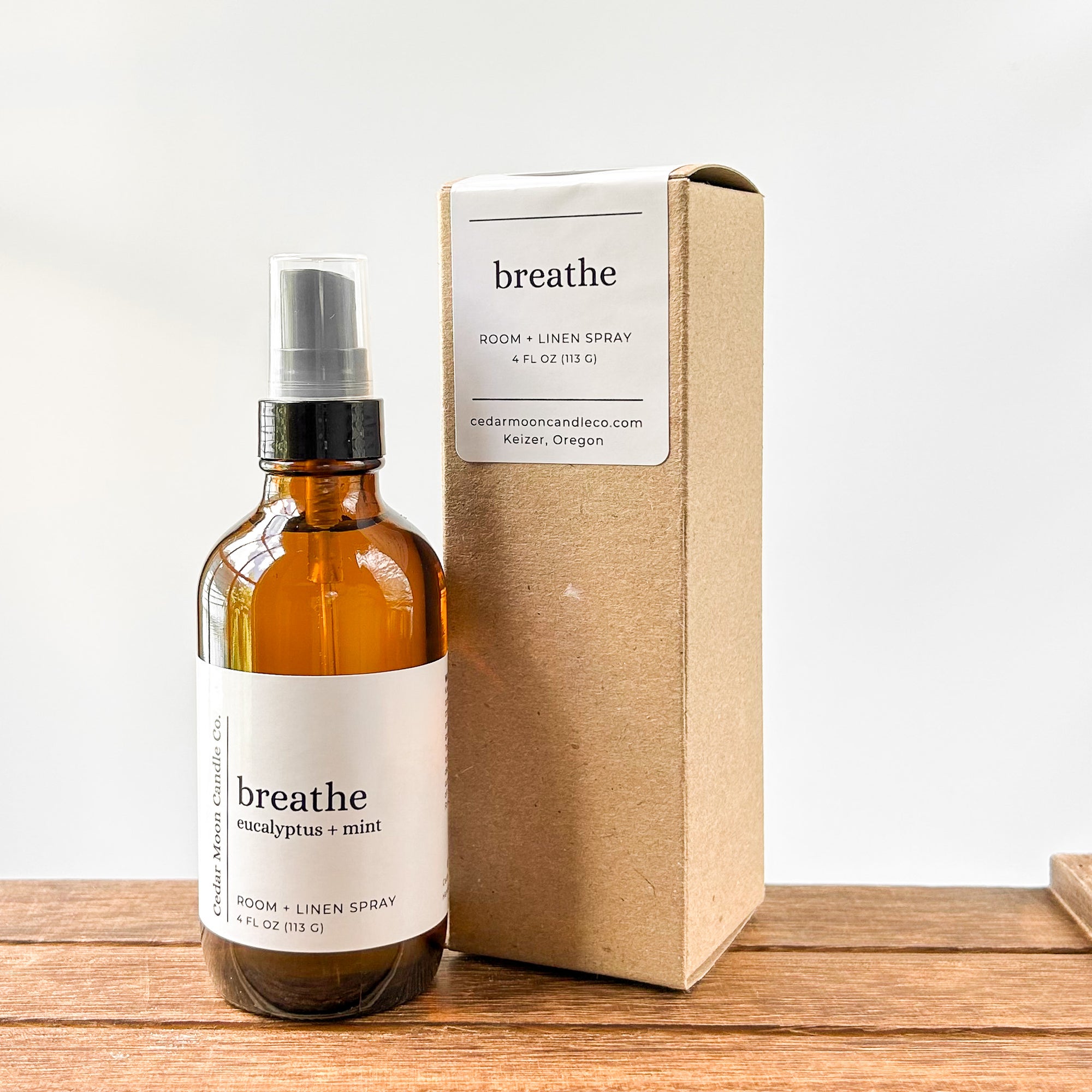 An amber glass bottle labeled 'breathe eucalyptus  mint' next to a brown cardboard box, both arranged on a wooden surface. The bottle is a room and linen spray, 4 fl oz.