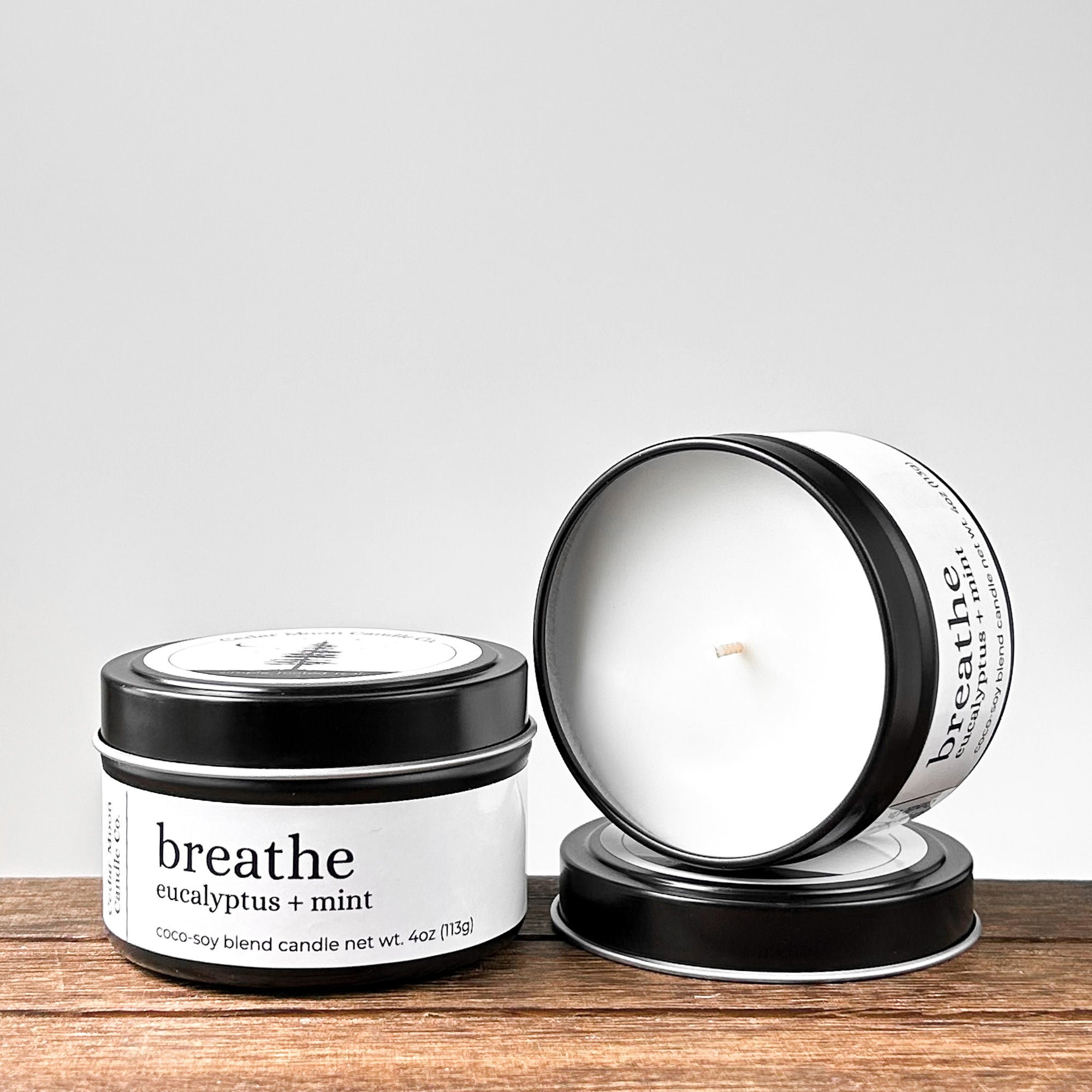 Two black candle tins labeled 'breathe eucalyptus  mint' with a wooden surface beneath, one lid off revealing the candle inside.