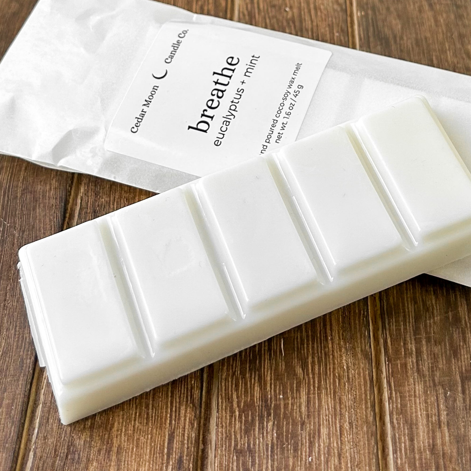 A white wax melt bar labeled 'breathe eucalyptus  mint' next to its packaging. The background features a wooden surface.