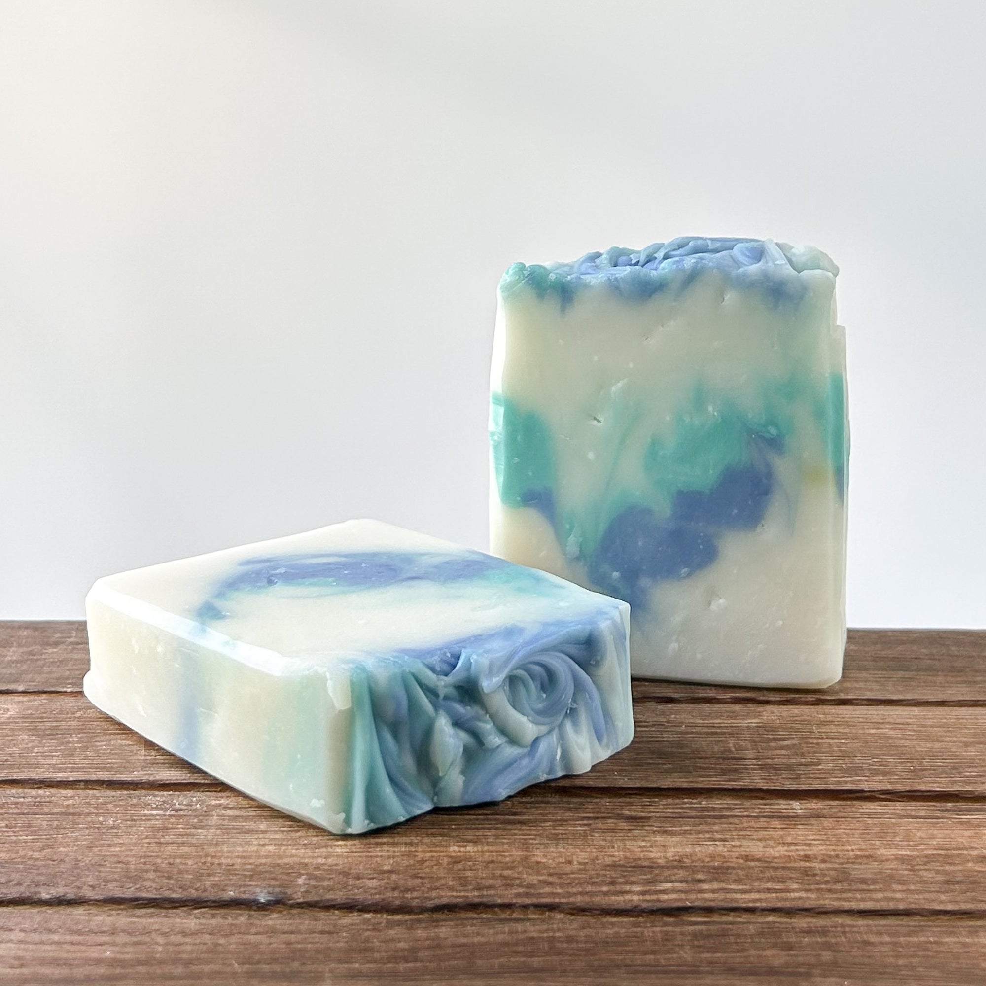 Two handmade soap bars with a swirl of blue and green colors on a wooden surface, against a light background.