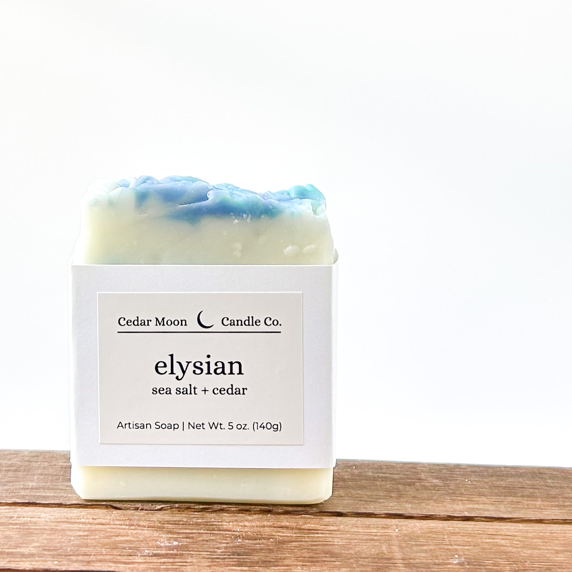 A bar of artisan soap with a blue and white top, labeled 'elysian sea salt  cedar'. The soap is resting on a wooden surface with a minimalist background.