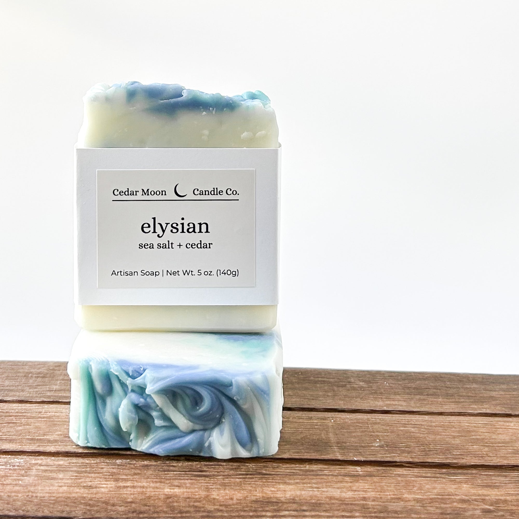 Two bars of artisan soap stacked on a wooden surface. The top bar is mainly white with a light blue swirl, while the bottom bar features a marbled blend of blue and white. Both bars have a label indicating 'elysian sea salt  cedar' from Cedar Moon Candle Co.