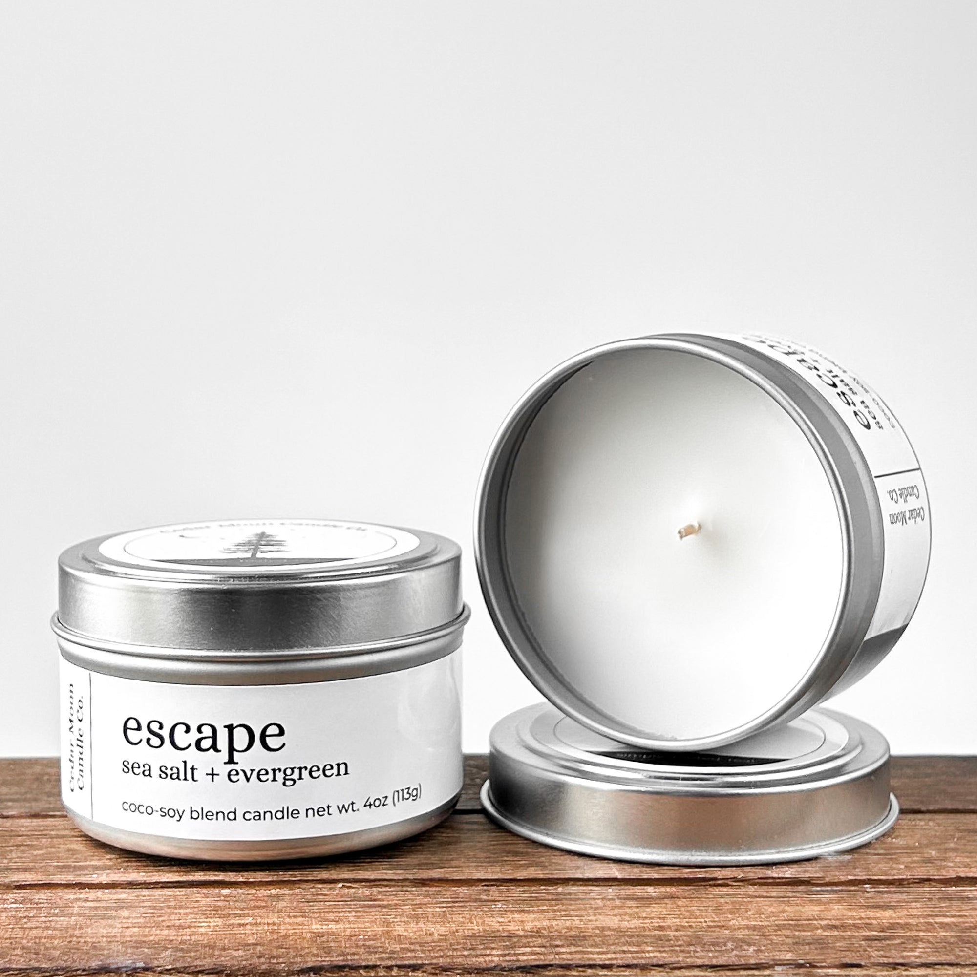 Two silver tins of 'escape sea salt  evergreen' coco-soy blend candle, one lid off, showcasing the white wax and wick. Placed on a wooden surface with a neutral background.
