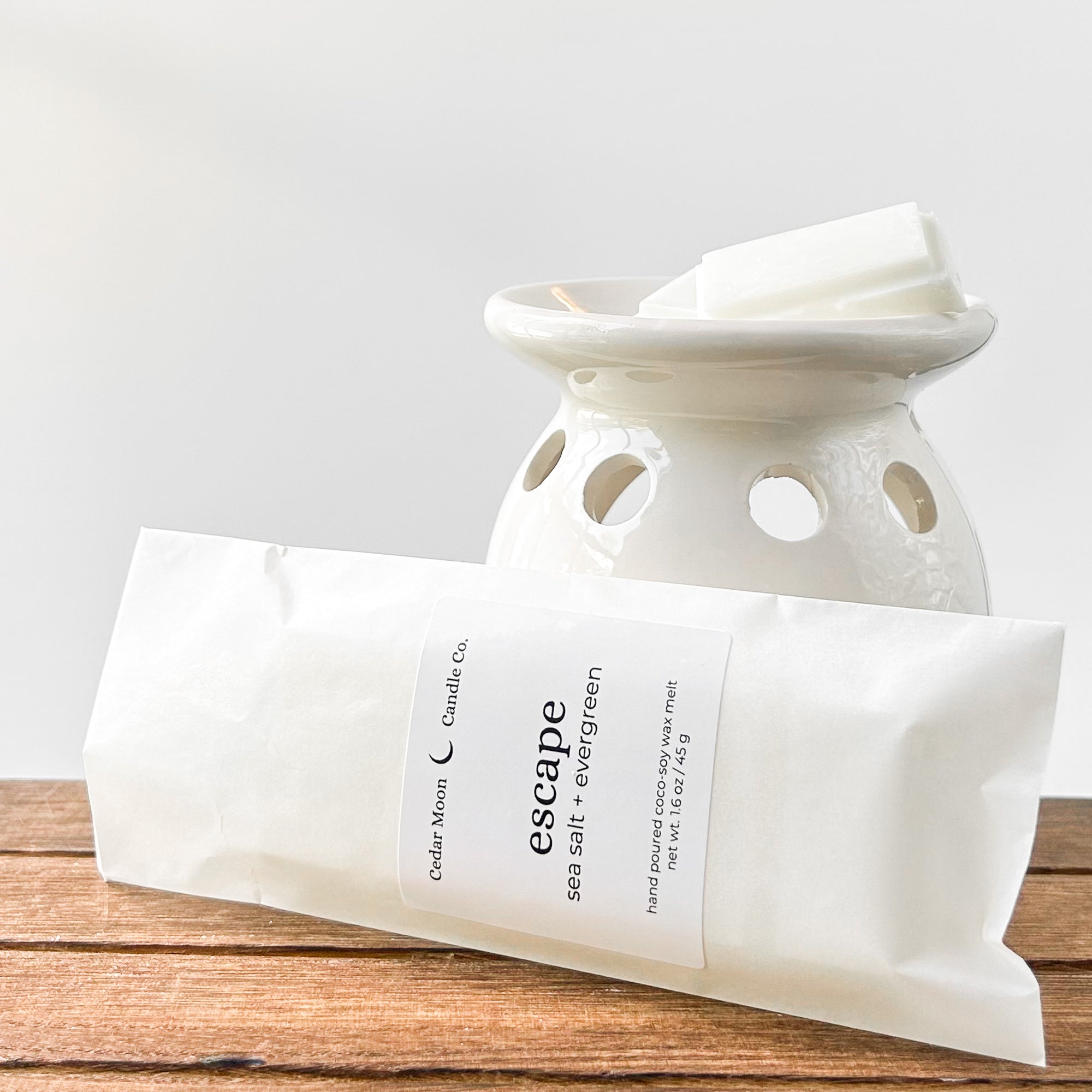 A ceramic wax warmer with white wax melts labeled 'escape' in sea salt and evergreen scent, placed on a wooden surface.
