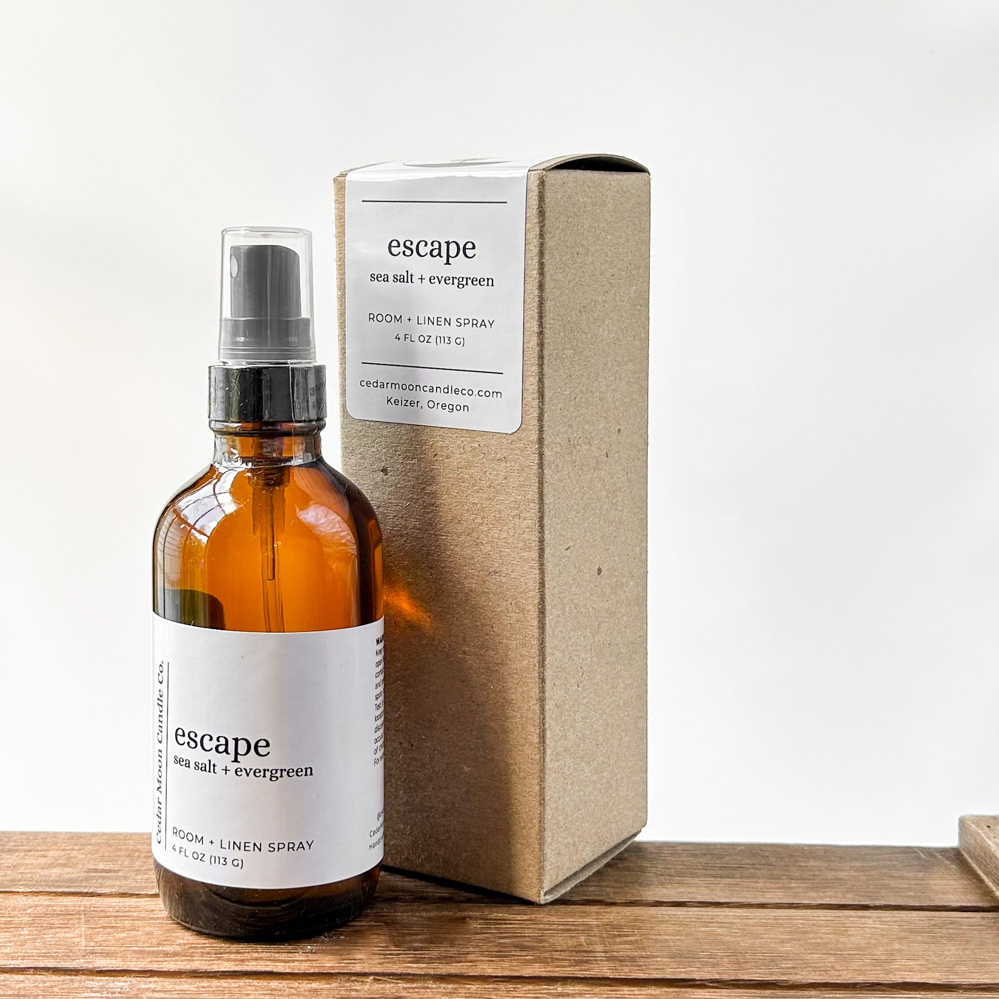A bottle of room and linen spray labeled 'escape sea salt  evergreen' next to a simple cardboard box, resting on a wooden surface with a neutral background.