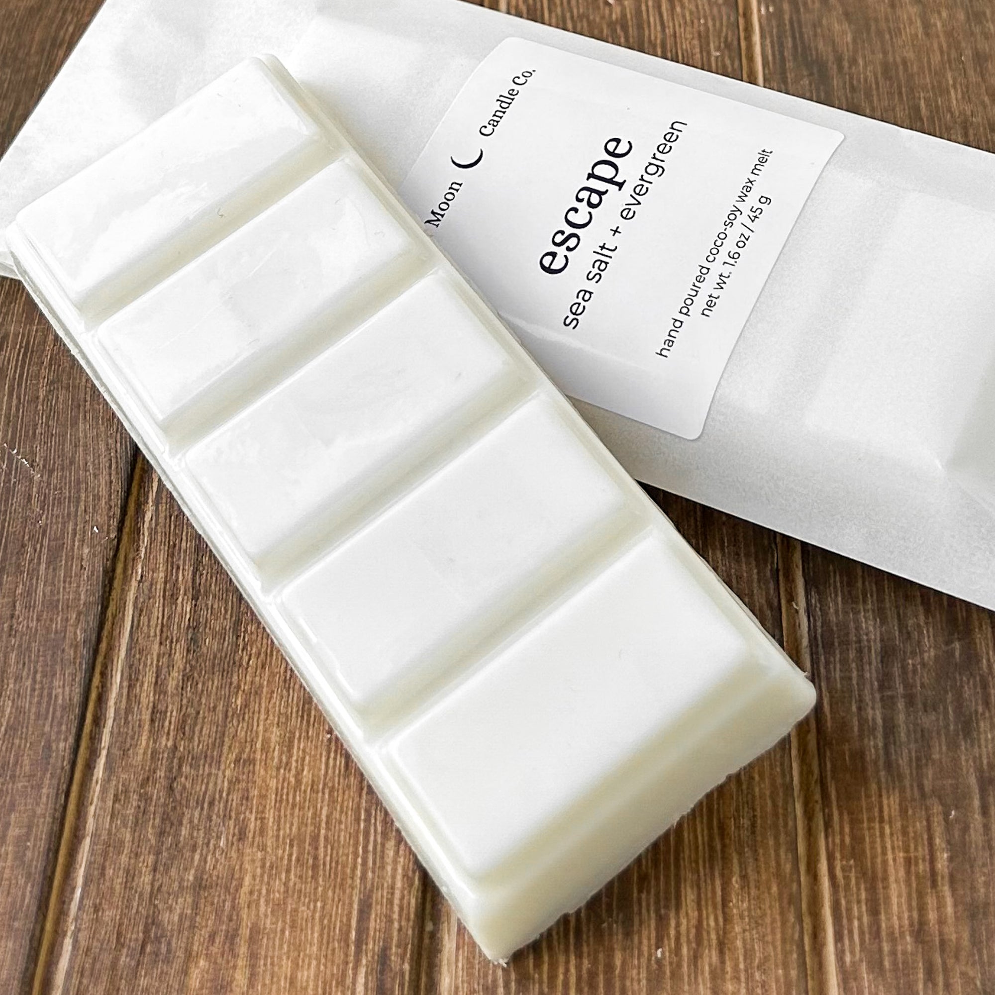 A white wax melt bar labeled 'escape' with scents of sea salt and evergreen, resting on a textured wooden surface. The package highlights the product's natural ingredients.