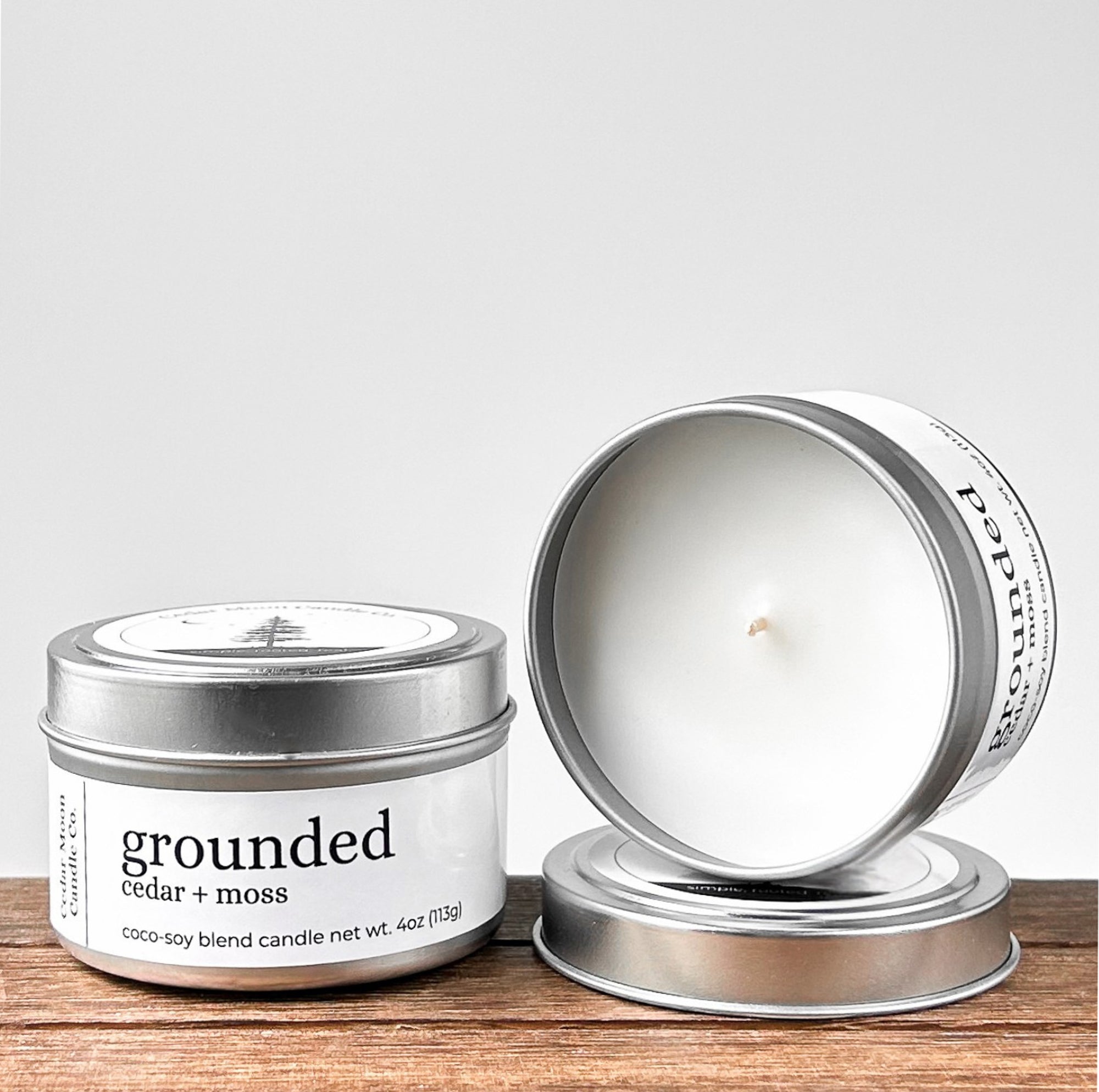Two silver tins of 'grounded cedar and moss' coco-soy blend candle, one lid off, showcasing the white wax and wick. Placed on a wooden surface with a neutral background.