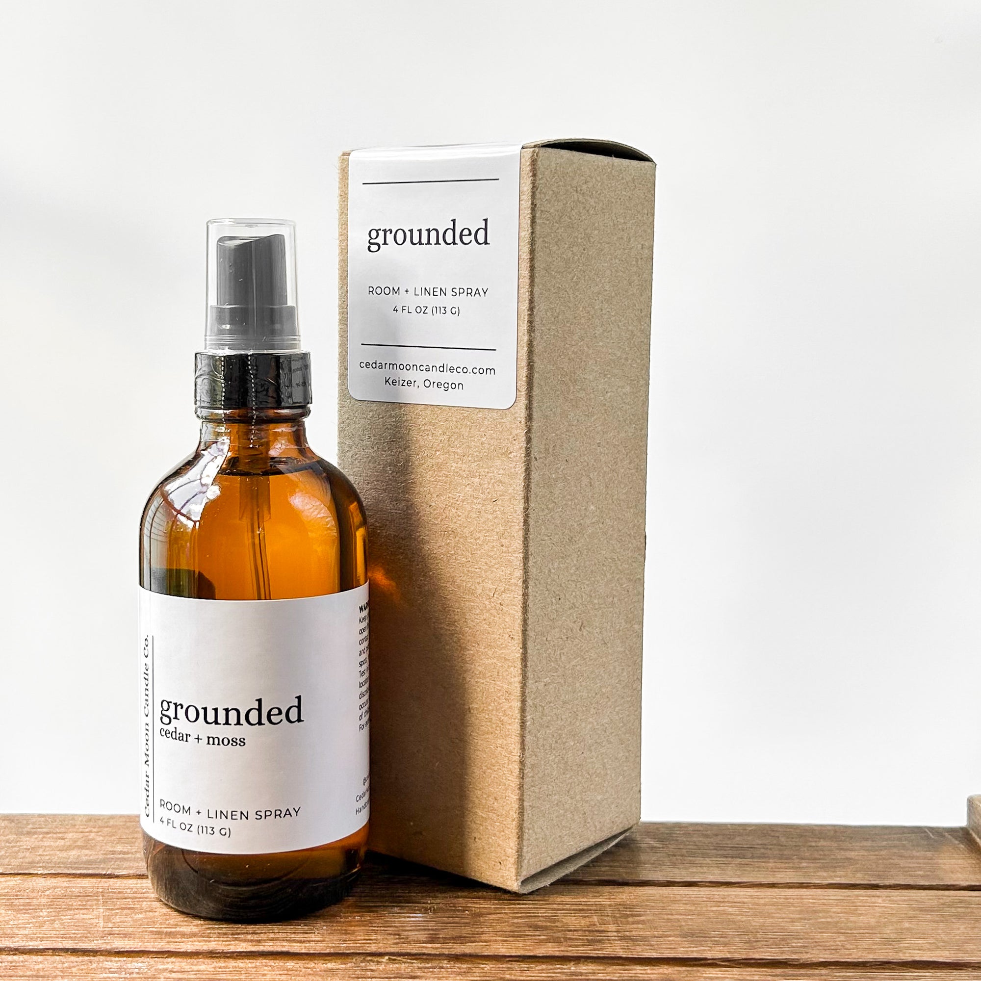 A bottle of room and linen spray labeled 'grounded cedar and moss' next to a simple cardboard box, resting on a wooden surface with a neutral background.