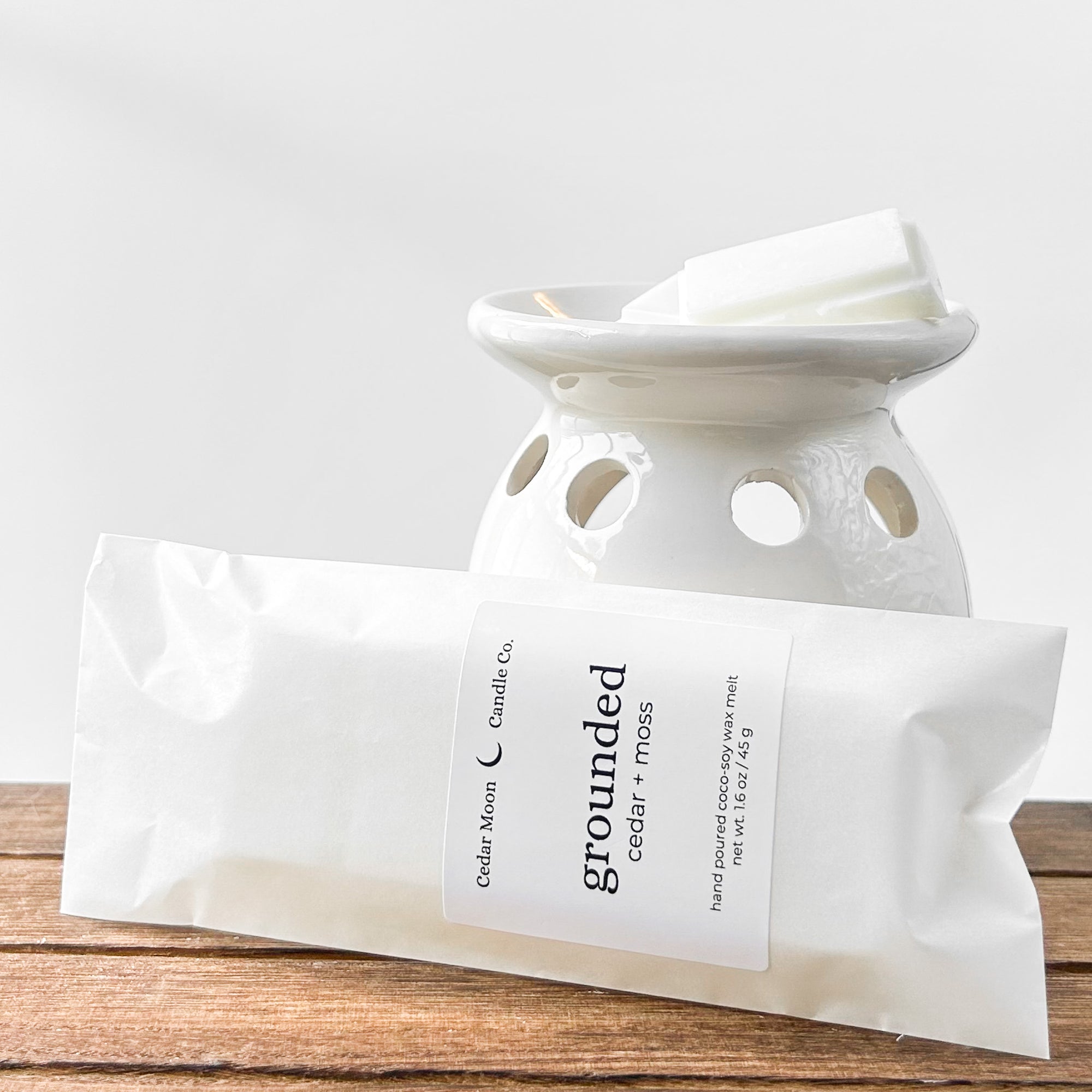 A ceramic wax warmer with white wax melts labeled 'grounded' in cedar and moss scent, placed on a wooden surface.