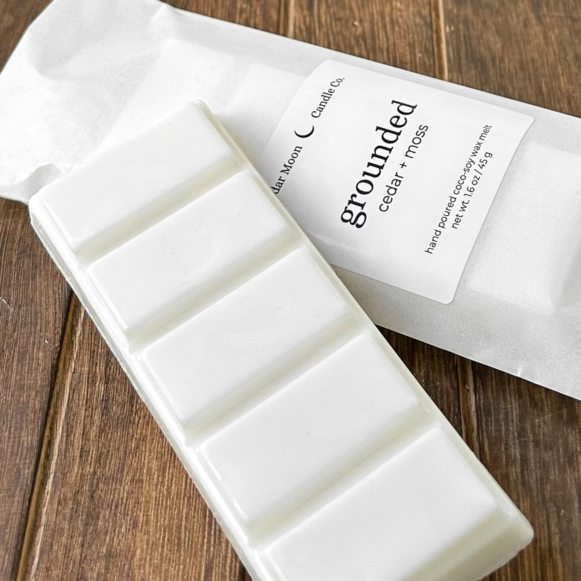 A white wax melt bar labeled 'grounded' with scents of cedar and moss', resting on a textured wooden surface. The package highlights the product's natural ingredients.