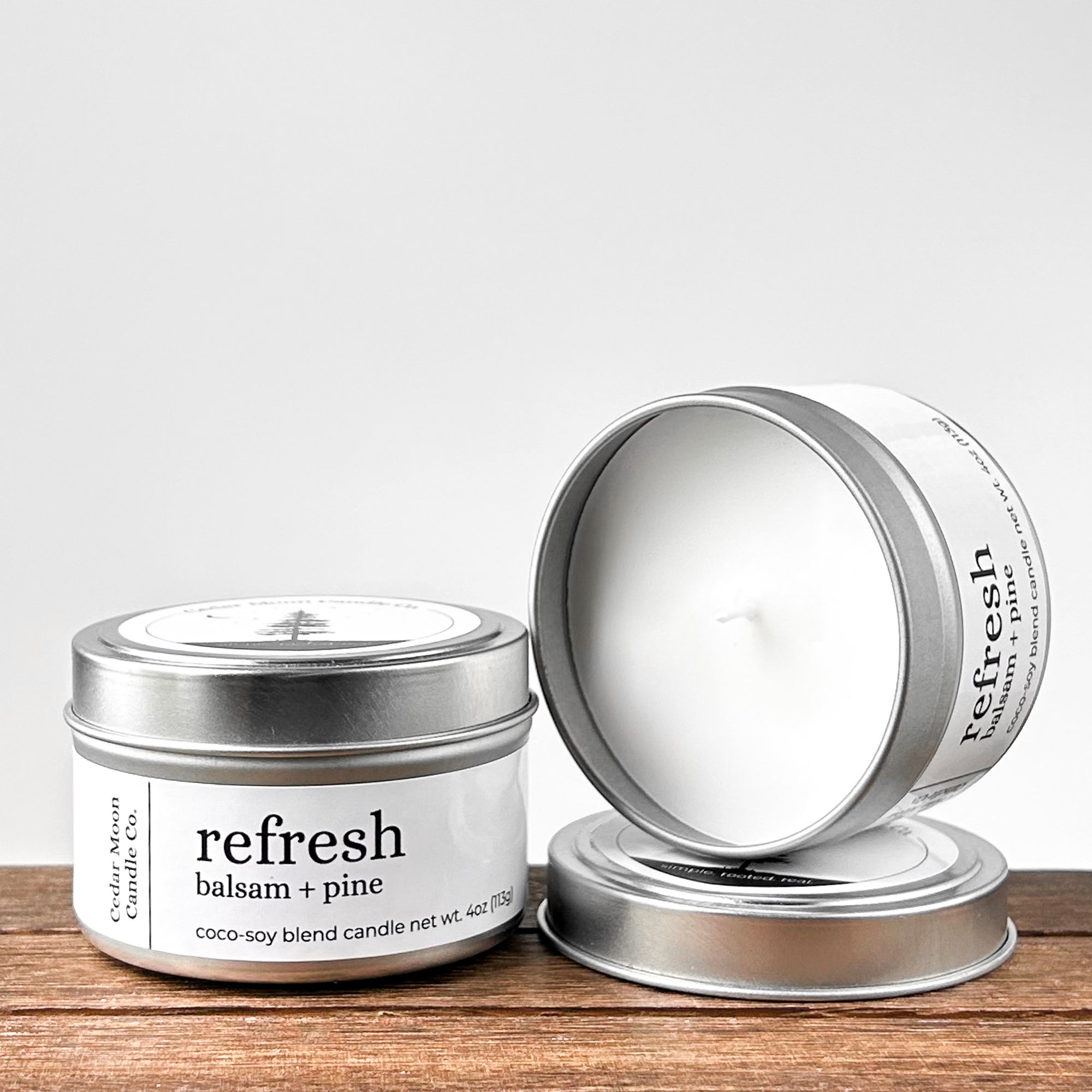 Two silver tins of 'refresh balsam and pine' coco-soy blend candle, one lid off, showcasing the white wax and wick. Placed on a wooden surface with a neutral background.