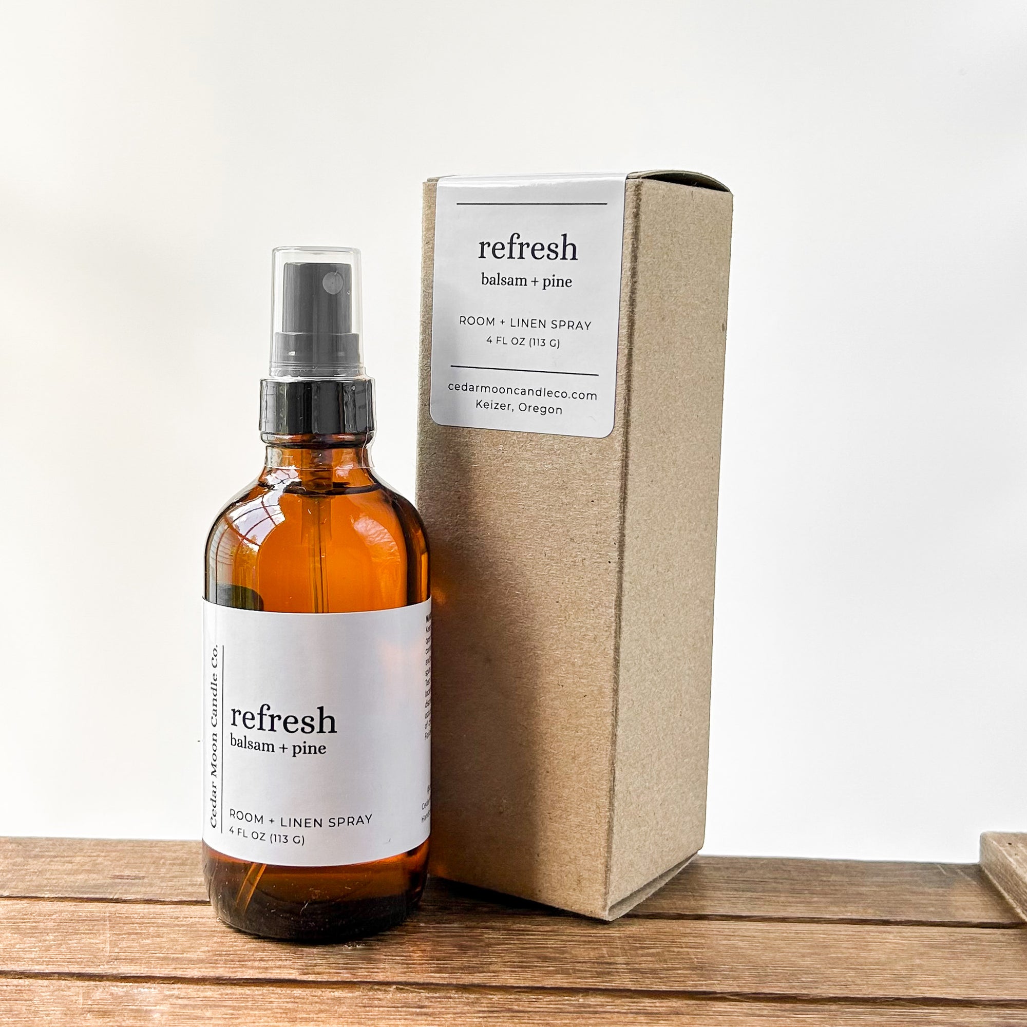 A bottle of room and linen spray labeled 'refresh balsam and pine' next to a simple cardboard box, resting on a wooden surface with a neutral background.