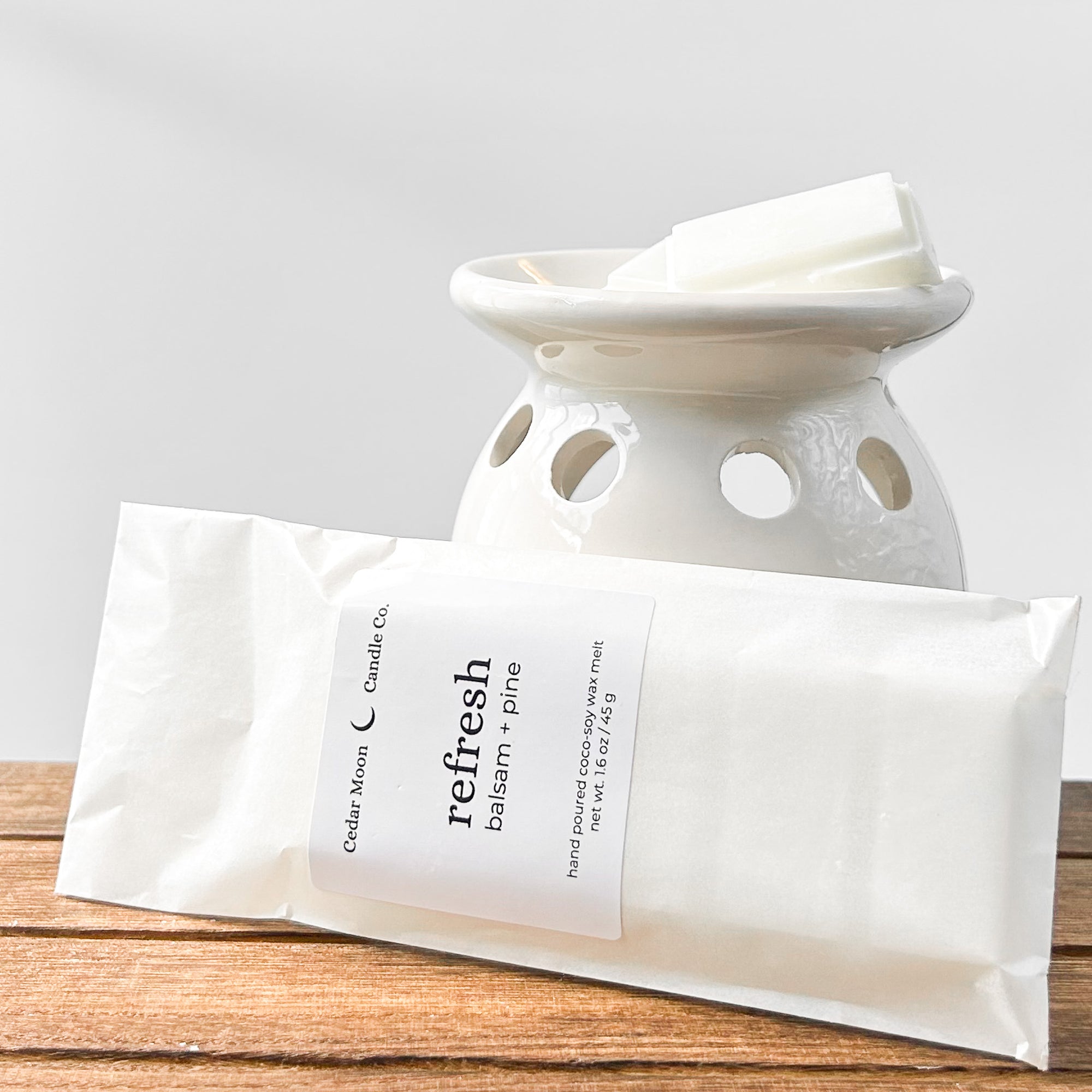 A ceramic wax warmer with white wax melts labeled 'refresh' in balsam and pine scent, placed on a wooden surface.