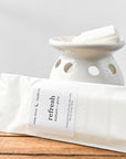 A ceramic wax warmer with white wax melts labeled 'refresh' in balsam and pine scent, placed on a wooden surface.