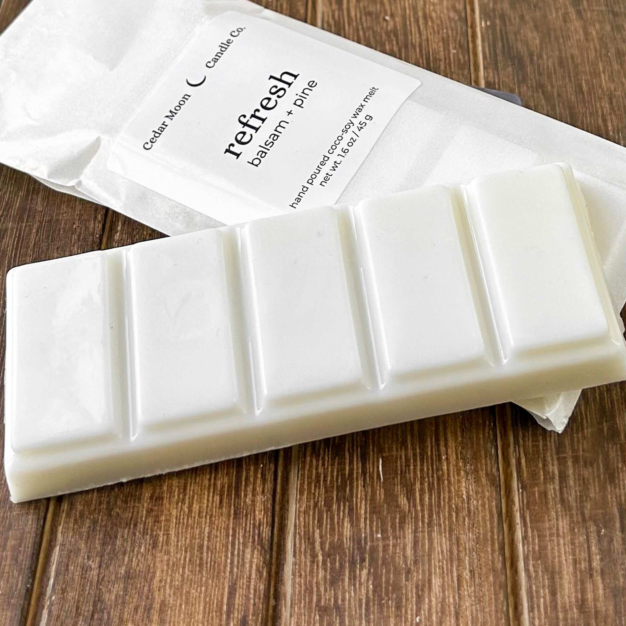 A white wax melt bar labeled 'refresh' with scents of balsam and pine, resting on a textured wooden surface. The package highlights the product's natural ingredients.