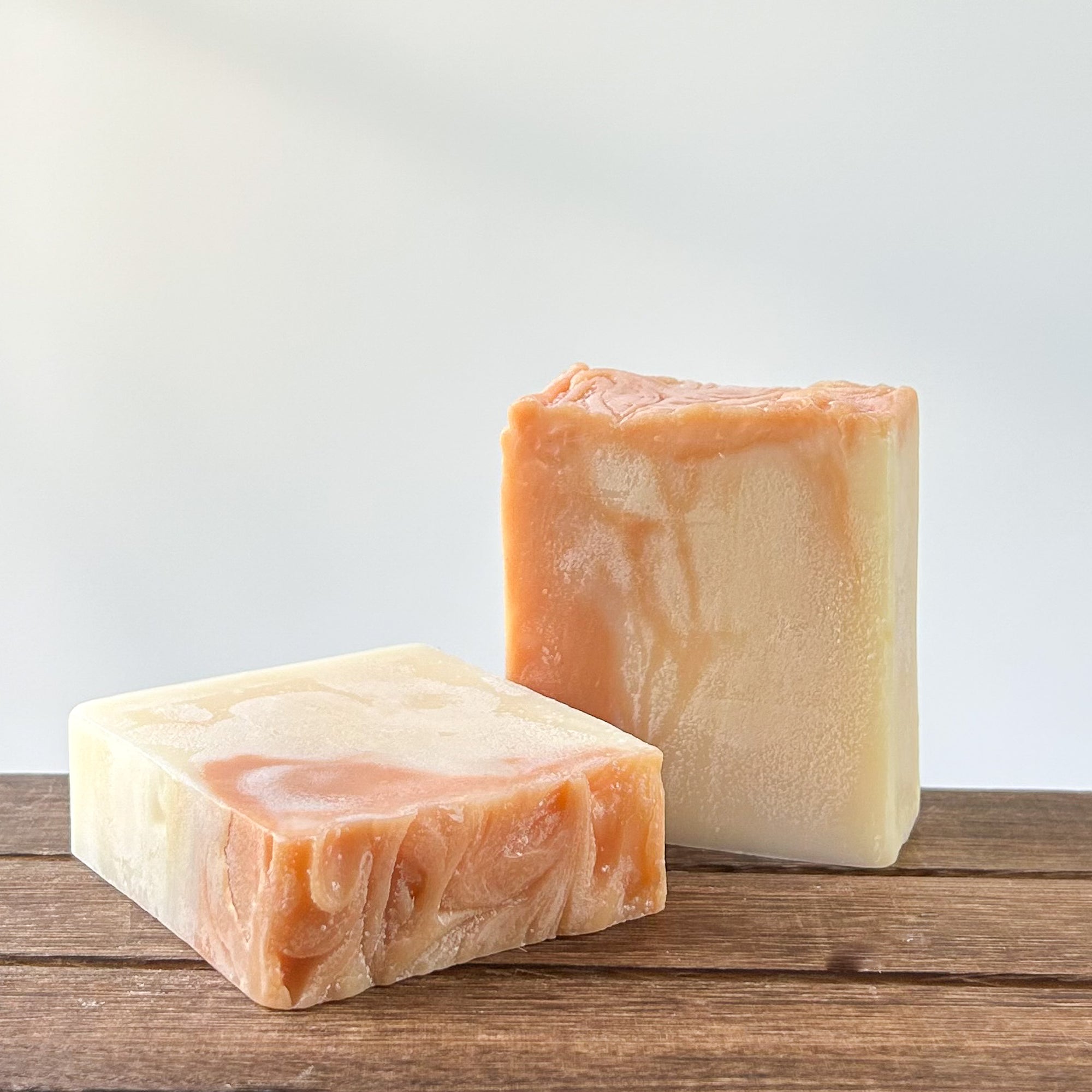 Two artisanal soap bars labeled 'renew' displayed on a wooden surface. One bar is cream-colored with swirling patterns, while the other has a light orange color. They are placed on a wooden surface with a light background.