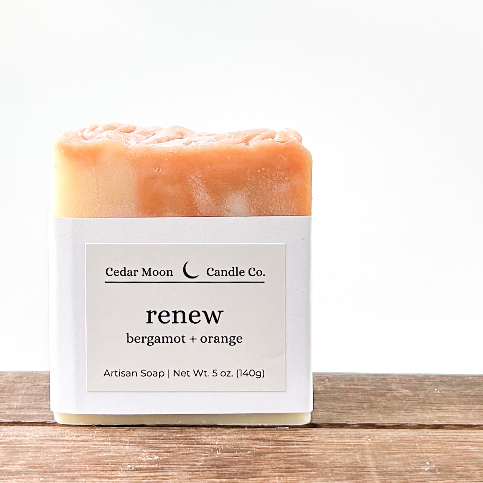 A bar of 'renew' artisan soap featuring bergamot and orange scents. The soap has a cream and orange swirl pattern and wrapped in a white paper band placed on a wooden surface against a light background.