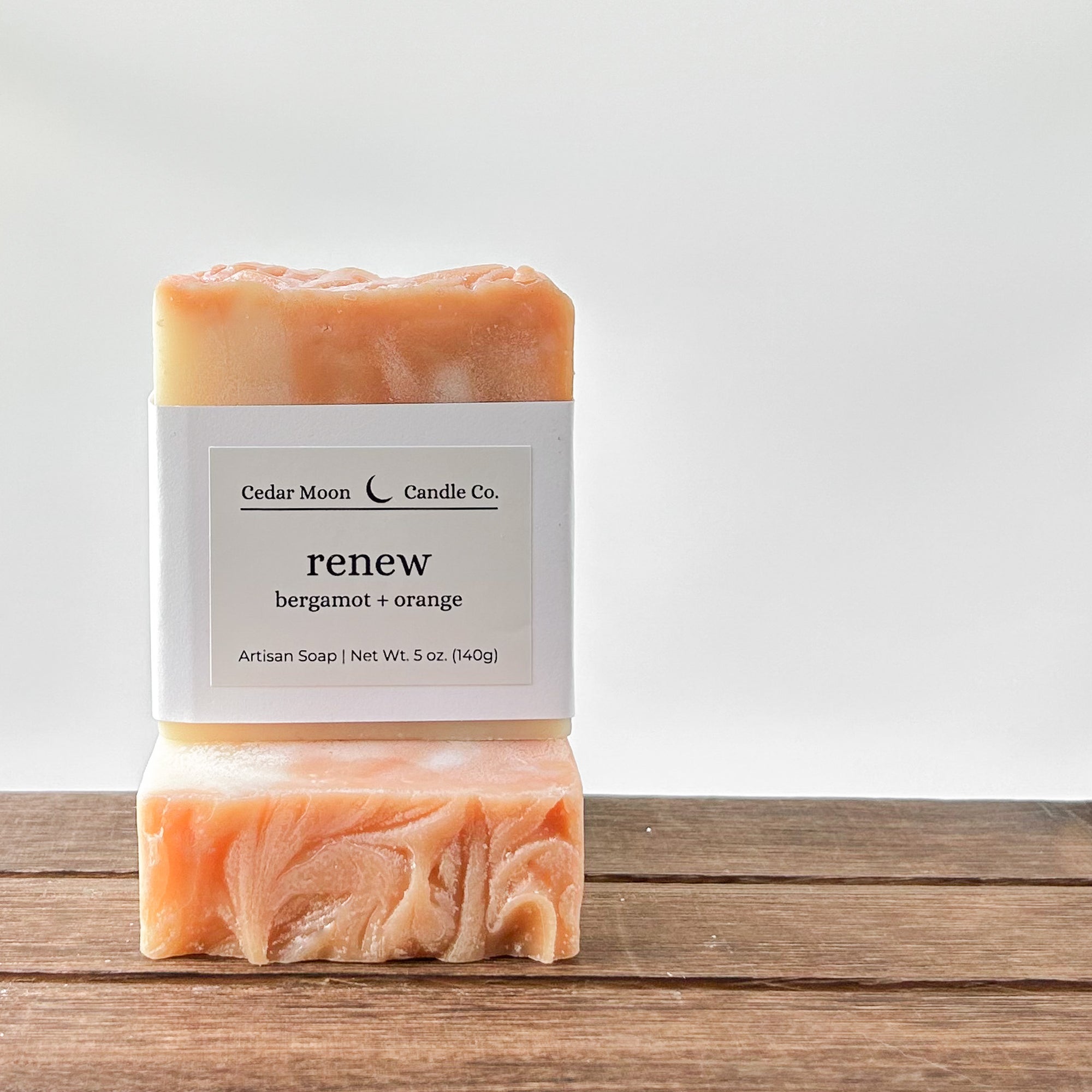 Two bars of artisan soap labeled 'renew' with bergamot and orange scent. The top bar has a smooth surface, while the bottom has a swirled design. They are placed on a wooden surface with a light background.